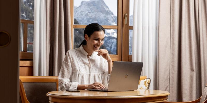 Donna sorridente lavora al laptop, vista montagna innevata dalla finestra, atmosfera accogliente.